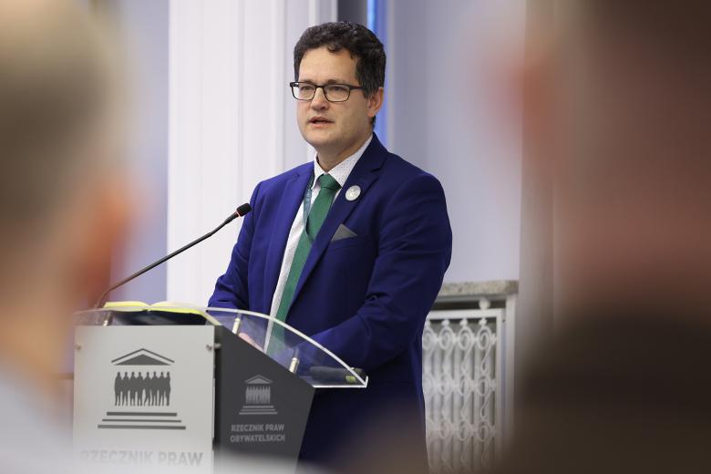 Mężczyzna w okularach i granatowym garniturze wypowiada się stojąc przy podium. 