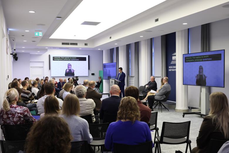 Widok ogólny sali konferencyjnej, na której siedzi kilkadziesiąt osób. Na środku mężczyzna w granatowym garniturze stoi przy mównicy i wypowiada się. Na ścianach są ekrany wyświetlające tłumaczkę języka migowego i napisy