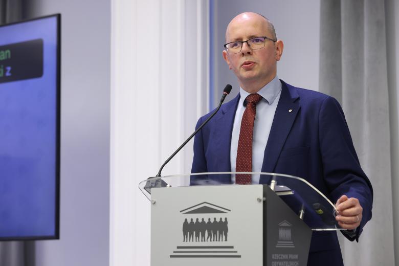 Mężczyzna w granatowym garniturze i bordowym krawacie wypowiada się stojąc przy mównicy