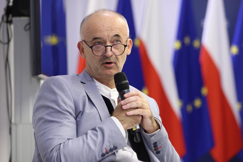 Mężczyzna w jasnoniebieskiej marynarce, okularach i koszuli bez kołnierzyka siedzi i wypowiada się do trzymanego w dłoniach mikrofonu