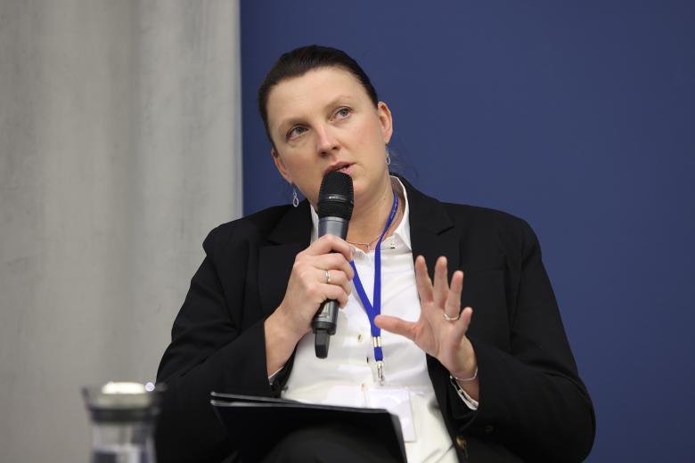 Kobieta w czarnej marynarce siedzi i wypowiada się do mikrofonu trzymanego w jednej dłoni, gestykulując drugą