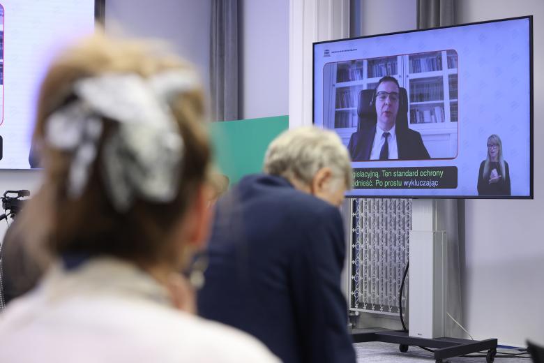 Ekran, na którym wyświetla się mężczyzna uczestniczący w panelu za pośrednictwem Internetu. Mężczyzna siedzi na tle regału z książkami i wypowiada się. Na pierwszym planie rozmyte głowy osób z widowni.