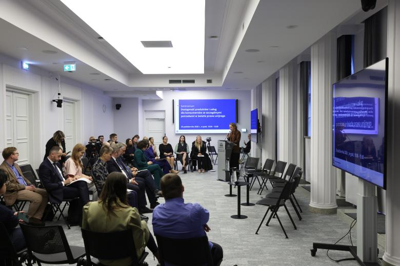 Sala konferencyjna, w której siedzi kilkadziesiąt osób. Na środku sali przy mównicy stoi kobieta i wypowiada się do mikrofonu. W szczycie sali jest ekran z napisem "Seminarium - Dostępność produktów i usług dla konsumentów ze szczególnymi potrzebami"