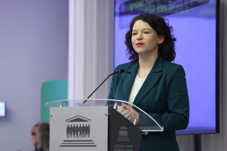 Kobieta w zielonej marynarce stoi przy mównicy i wypowiada się do mikrofonu
