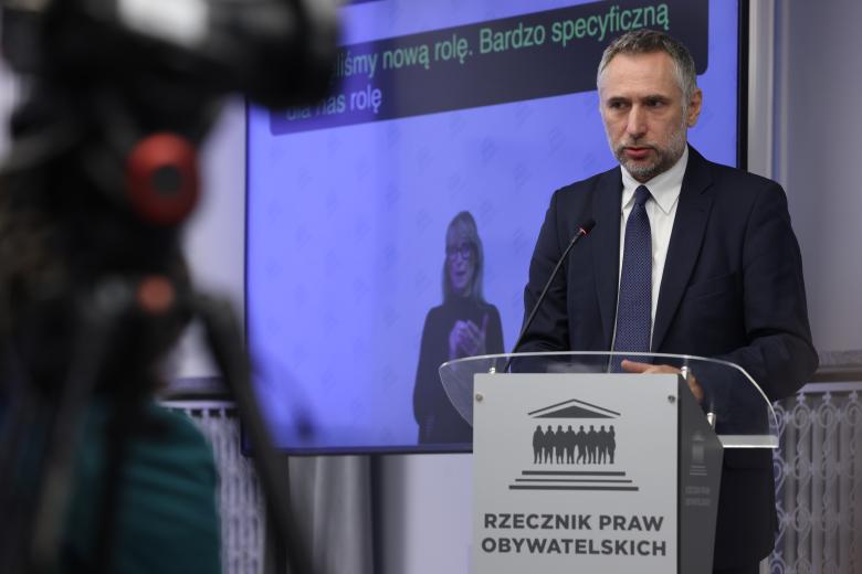 Mężczyzna z krótką brodą w granatowym garniturze wypowiada się stojąc przy mównicy z mikrofonem. Za nim jest ekran, na którym wyświetla się tłumaczka języka migowego i napisy