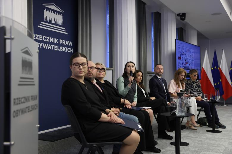 Osiem osób siedzi na krzesłach ustawionych w szeregu i spogląda w jedną stronę. Jedna z nich mówi do trzymanego w dłoni mikrofonu. 