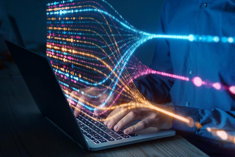 Osoba pisze na klawiaturze laptopa, ponad klawiaturą unosi się świecąca wizualizacja przepływu danych komputerowych