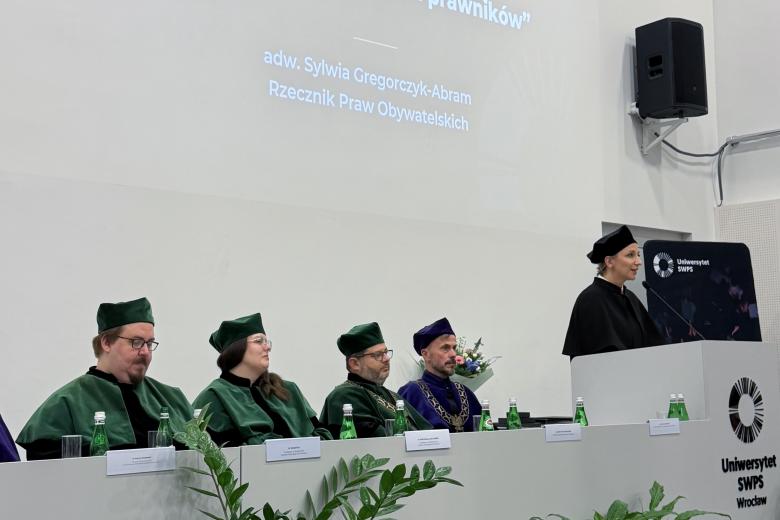 Cztery osoby w togach siedzą przy stole w szczycie auli akademickiej. Obok nich przy podium stoi Sylwia Gregorczyk-Abram i wypowiada się do mikrofonu