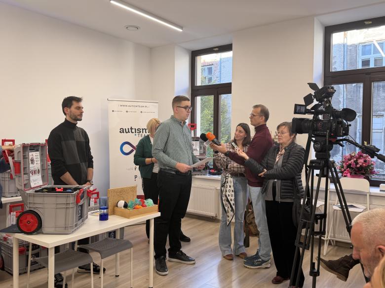 Mężczyzna wypowiada się do mikrofonów trzymanych przez trzech dziennikarzy. Obok niego jest stół, na którym stoi skrzynka sensoryczna, a za nim stoją dwie osoby.  