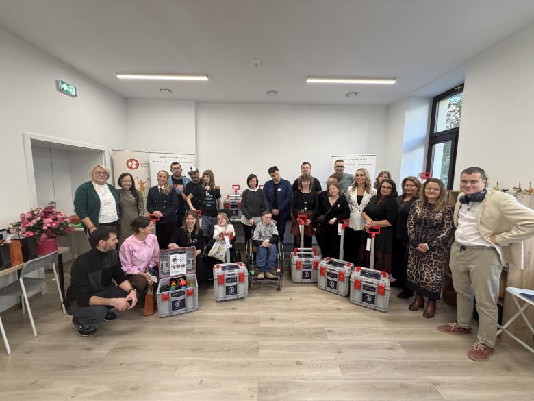 Zdjęcie grupowe ok. 20 osób stojących w pomieszczeniu. Przed nimi stoi 5 skrzyneczek pomocy sensorycznej