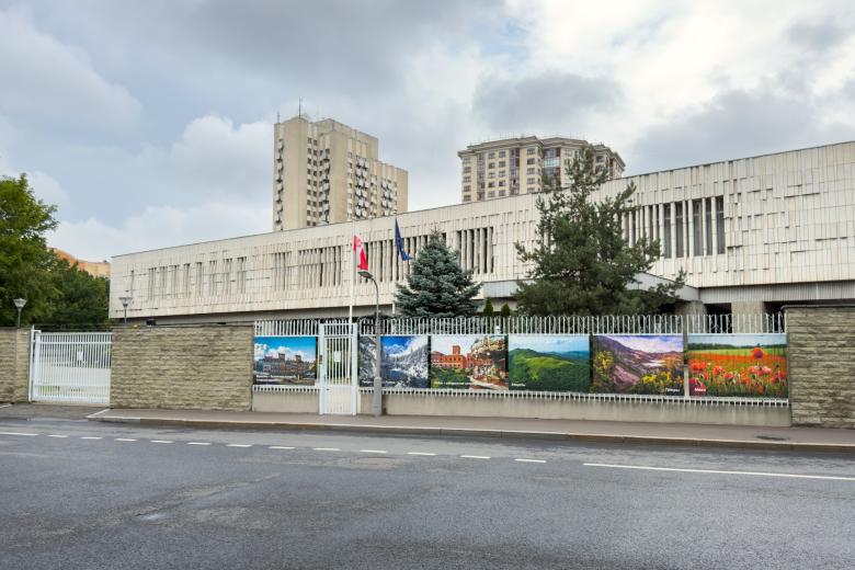 Kilkupiętrowy długi budynek widziany z boku otoczony ogrodzeniem, przed budynkiem na masztach flagi Polski i UE