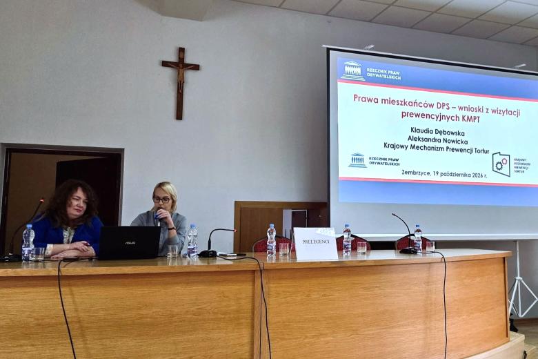 Dwie osoby siedzą za dużym stołem na sali konferencyjnej, jedna z nich trzyma mikrofon, za nimi w tle na wyświetlaczu widoczna prezentacja