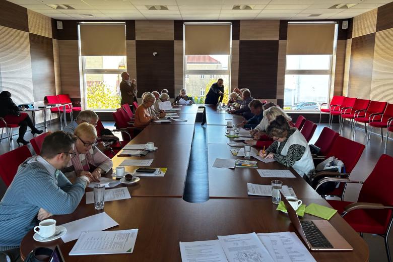 Osoby starsze siedzą na krzesłach w dużej sali, są pochylone nad stołem, wypełniają zadania warsztatowe