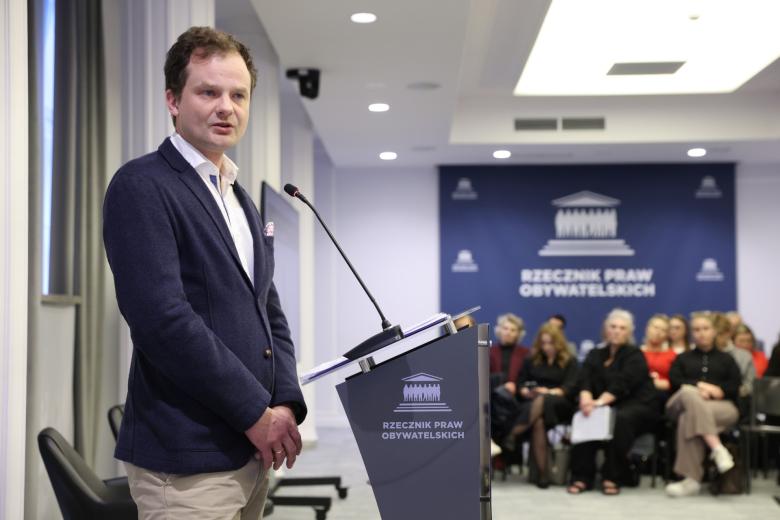 Osoba przemawia za mównicą na sali konferencyjnej wypełnionej ludźmi
