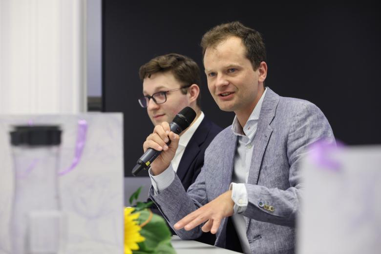 Osoba siedzi za stołem w sali konferencyjnej i mówi do trzymanego w dłoni mikrofonu