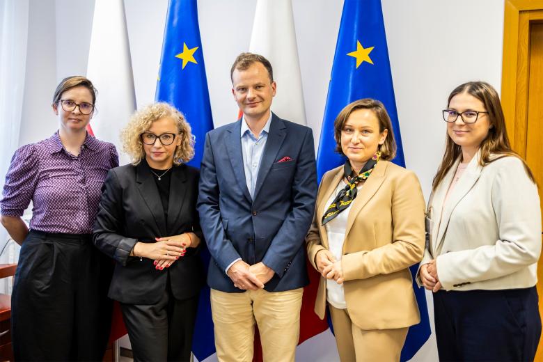 Pięć osób pozuje do wspólnego zdjęcia na tle flag Polski i UE