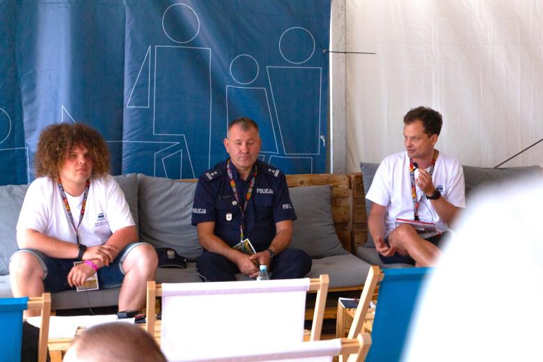 Trzy osoby siedza na kanapach w duży namiocie festiwalowym, jedna z nich mówi do trzymanego w dłoni mikrofonu