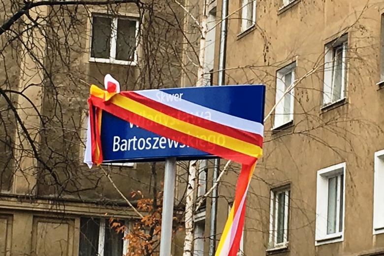 Zdjęcie: tablica informacyjna "Skwer Władysława Bartoszewskiego" przyslonięta jeszcze barwami narodowymi i żółto-czerwonymi barwami Warszawy