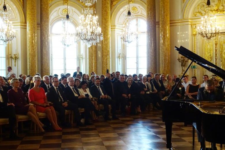 zdjęcie: po prawej stronie widać fragment fortepiany i grającego na nim mężczyznę, w tle widać kilkadziesiąt siedzących osób