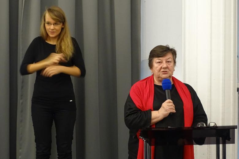 zdjęcie: kobieta w czarnej sukience i czerwonym szalu stoi przy mównicy, za nią kobieta ubrana na czarno, która gestykuluje