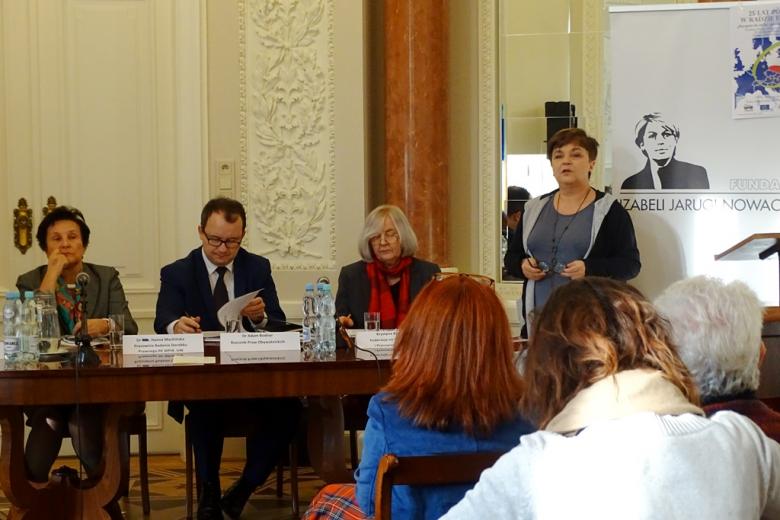 zdjęcie: kobieta stoi przy mównicy po jej lewej stronie za stołem siedzą dwie kobiety i mężczyzna
