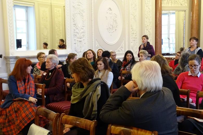 zdjęcie: kilkanaście osób siedzi na sali, jedna z kobiet mówi do mikrofonu