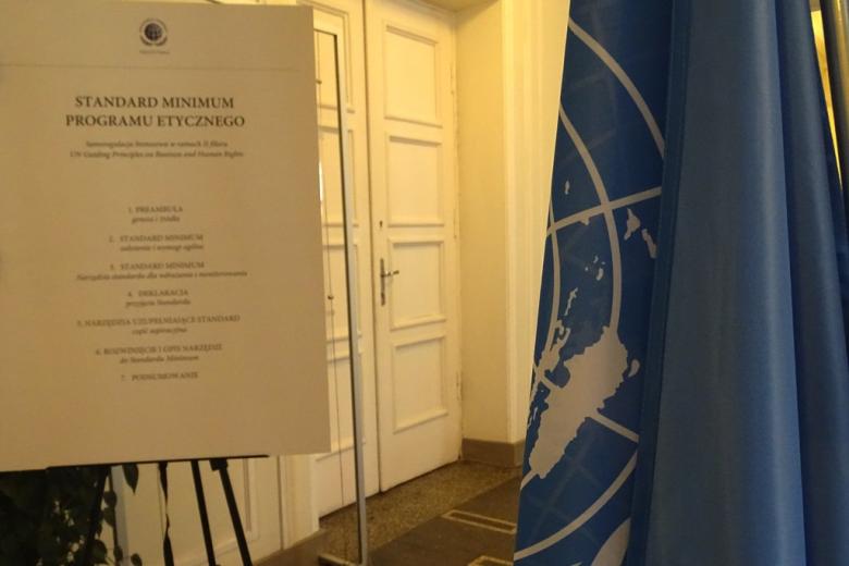zdjęcie: na pierwszym planie fragment flagi ONZ w tle tablica z opisen Standardów Minimum Programu Etycznego