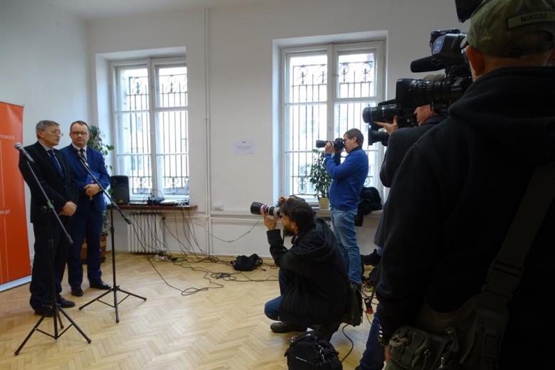 zdjęcie: po prawej stronie widać kilkunastu dziennikarzy z aparatami i kamerami, po lewej stronie stoi dwóch mężczyzn w garniturach