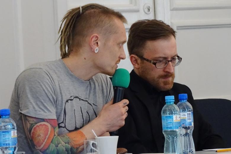 zdjęcie: przy stole siedzi dwóch mężczyn, jeden z nich z tatuażem na ręku mówi do mikrofonu