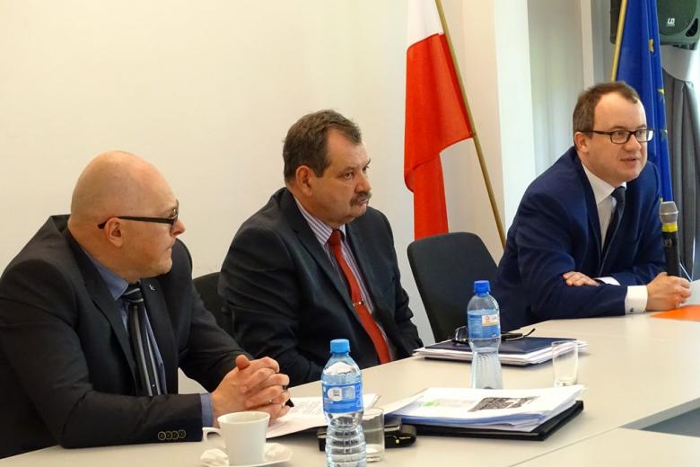 zdjęcie: trzej mężczyźni siedzą przy stole konferencyjnym, za mężczyzną po prawej stronie widać flegi Polski i Unii Europejskiej