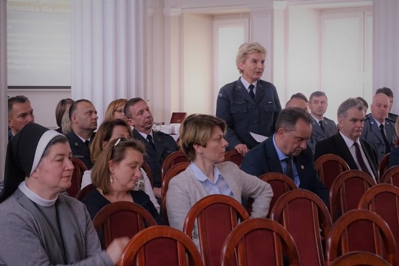 zdjęcie: kilka osób siedzi na sali, kobieta w munduże stoi pośrodku nich