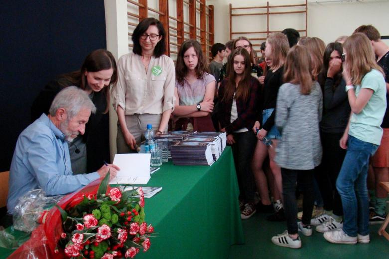 zdjęcie: po lewej stronie stół przy którym siedzi mężczyzna i podpisuje książki, obok niego stoi kobieta, za nią kilkudziesięciu uczniów
