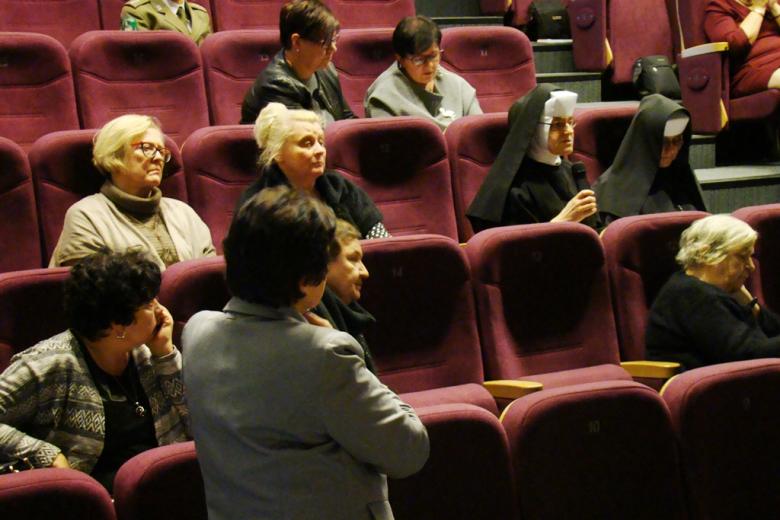 zdjęcie: kilka osób siedzi na sali, jedna z kobiet w habicie mówi do mikrofonu, przed nią stoi kobieta w marynarce