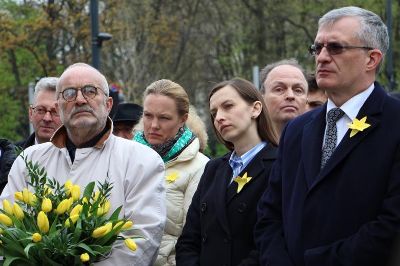 zdjęcie: mężczyzna w jasnym płaszczu trzyma w rękach bukiet żółtych tulipanów, obok niego stoi kobieta i mężczyzna w ciemnych płaszczach