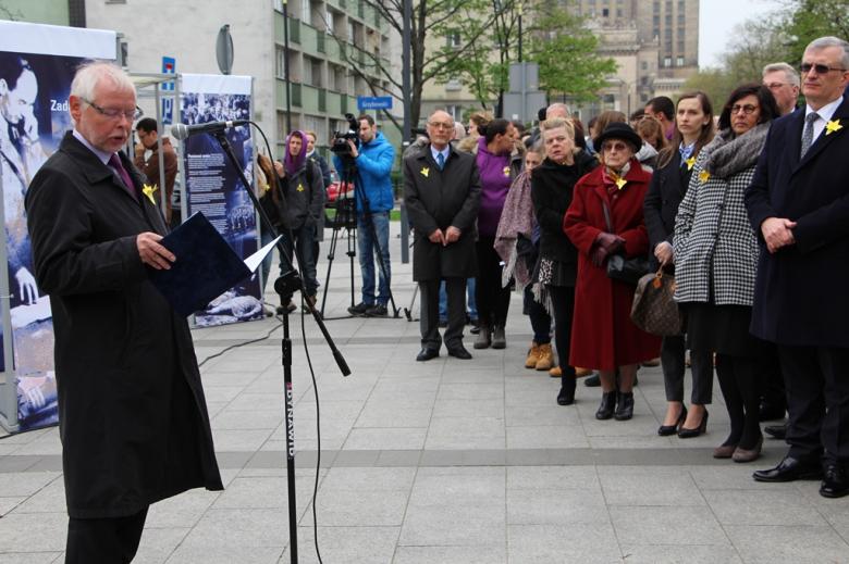 zdjęcie: po lewej stronie stoi mężczyzna w czarnym płaszczu i mówi do mikrofonu, za nim widać fragmenty niebieskich plansz, po prawej stronie stoi grupa osób