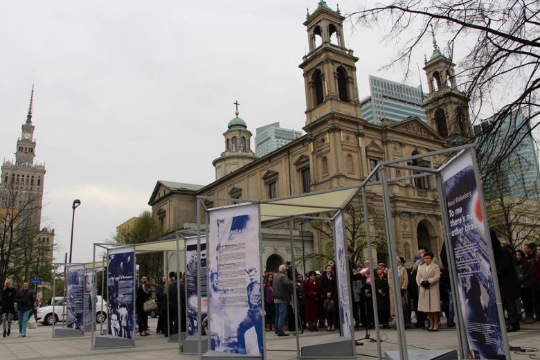 zdjęcie: na pierwszym planie widać niebiesko-białe plansze, za nimi stoją uczestnicy uroczystości w tle widać kościół, a po lewej stronie Pałac Kultury i Nauki