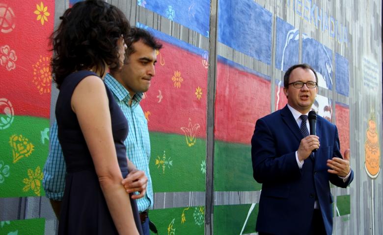 zdjęcie: na tle muralu w niebiesko-czerwono-zielonych barwach, stoją dwaj mężczyźni i jedna kobieta, mężczyna po prawej - w granatowym garniturze przemawia do mikrofonu