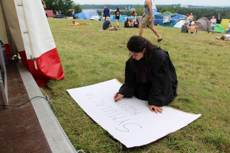 zdjęcie: kobieta w czarnej todze kłeczy na trawie i rysuje plakat