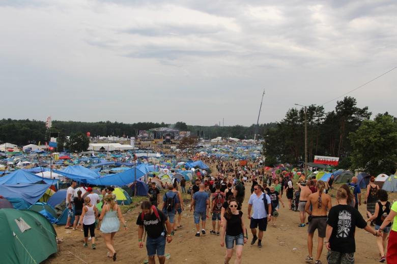zdjęcie: na pierwszy planie pole namiotowe i idący ludzie, w tle ogromna scena