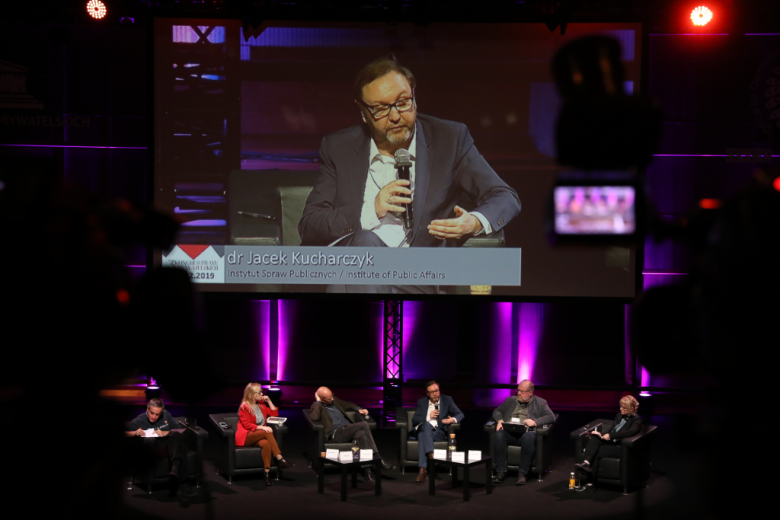 Paneliści na scenie, na ekranie za nimi powiększona postać z podpisem Jacek Kucharczykj