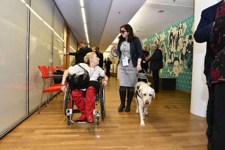 Kobieta z psem asystującym i kobieta na wózku idą korytarzem