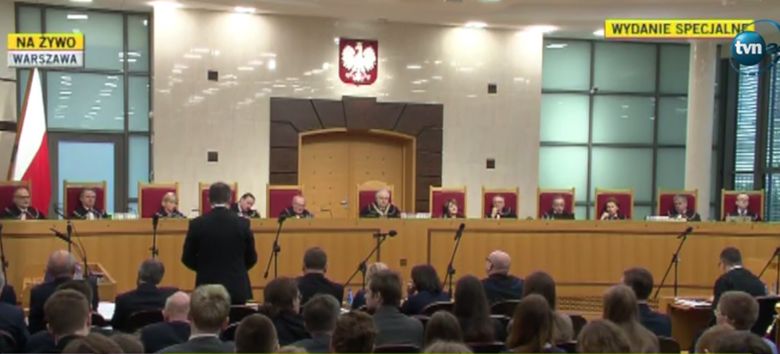 Zdjęcie bardzo szerokie, widac pełen skłąd Trybunału: sędziowie siedzą, z lewej strony męzczyzna widziany od tyłu przemawia na stojąco