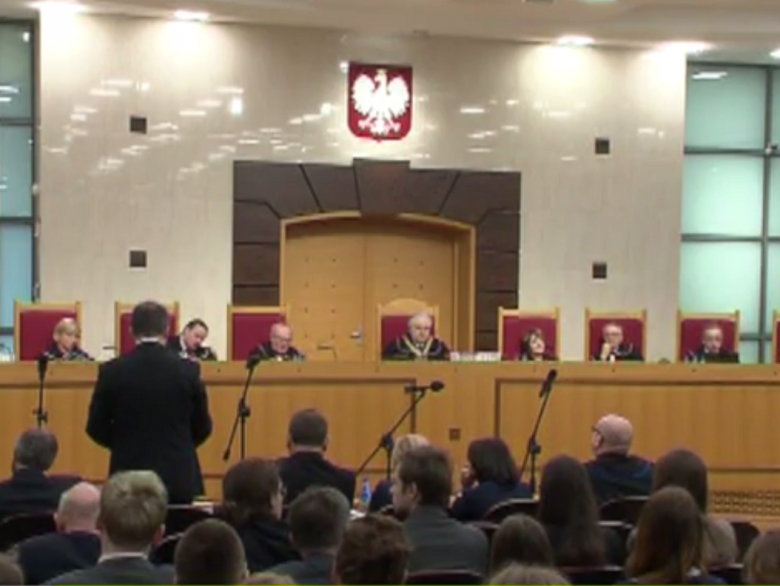 Zdjęcie: sędziowie siedzą, z lewej strony męzczyzna widziany od tyłu przemawia na stojąco