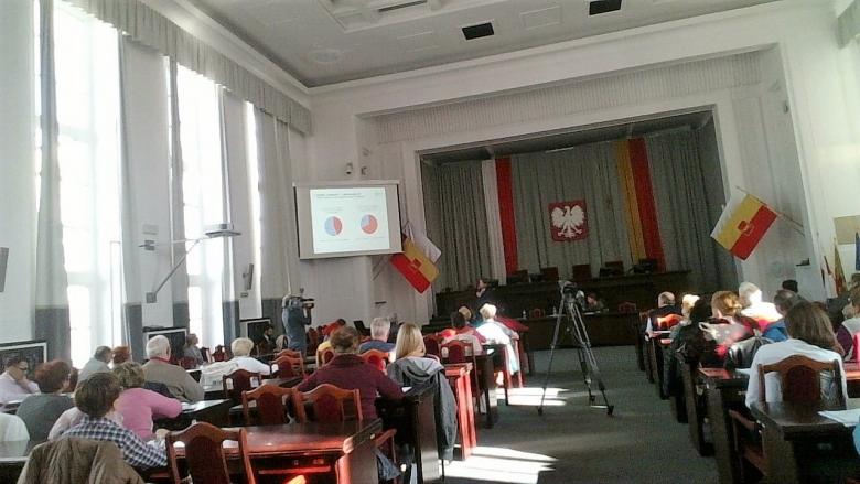 Ludzie siedzą  na sali obrad władz samorządowych. Flagi Łodzi