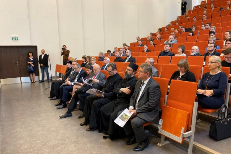 Ludzie na sali audytoryjnej, w tym ksiądz prawosławny, szersze ujęćie