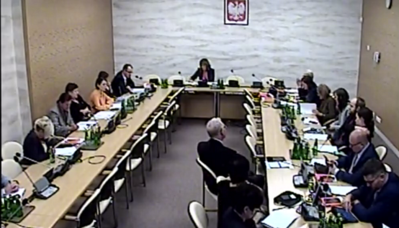 Screen komputerowy: posiedzenie komisji - ludzie siedza na sali