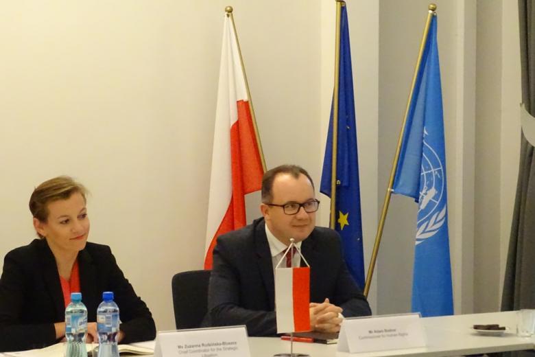 Kobieta i mężczyzna przy stole, z tyłu flagi Polski, UE i Narodów Zjednoczonych