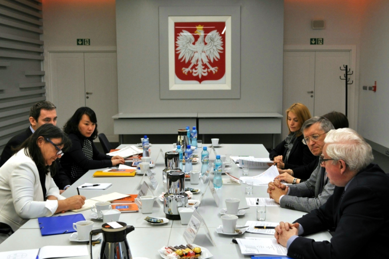 zdjęcie: po dwóch stronach stołu siedzi na przeciwko siebie sześć osób w tle na ścianie wisi godło Polski