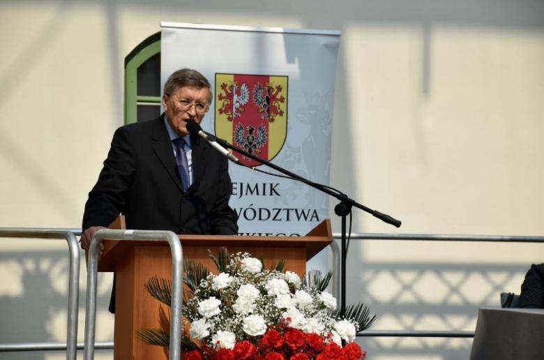 zdjęcie: mężczyzna w garniturze stoi przy mównicy, przed nią biało-czerwone kwiaty
