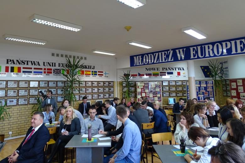 Zdjęcie: młodzi ludzie przy stolikach w sali bibliotecznej piszą na kartkach. Na ścianie napis "Młodzież w Europie" i lista krajów członkowskich UE
